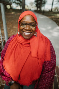 A woman sitting outside, smiling at the camera wearing an orange headscarf, square black glasses, and a pink shirt. 