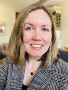 A close-up of a woman with short light brown hair smiling at the camera, wearing a brown plaid blazer.